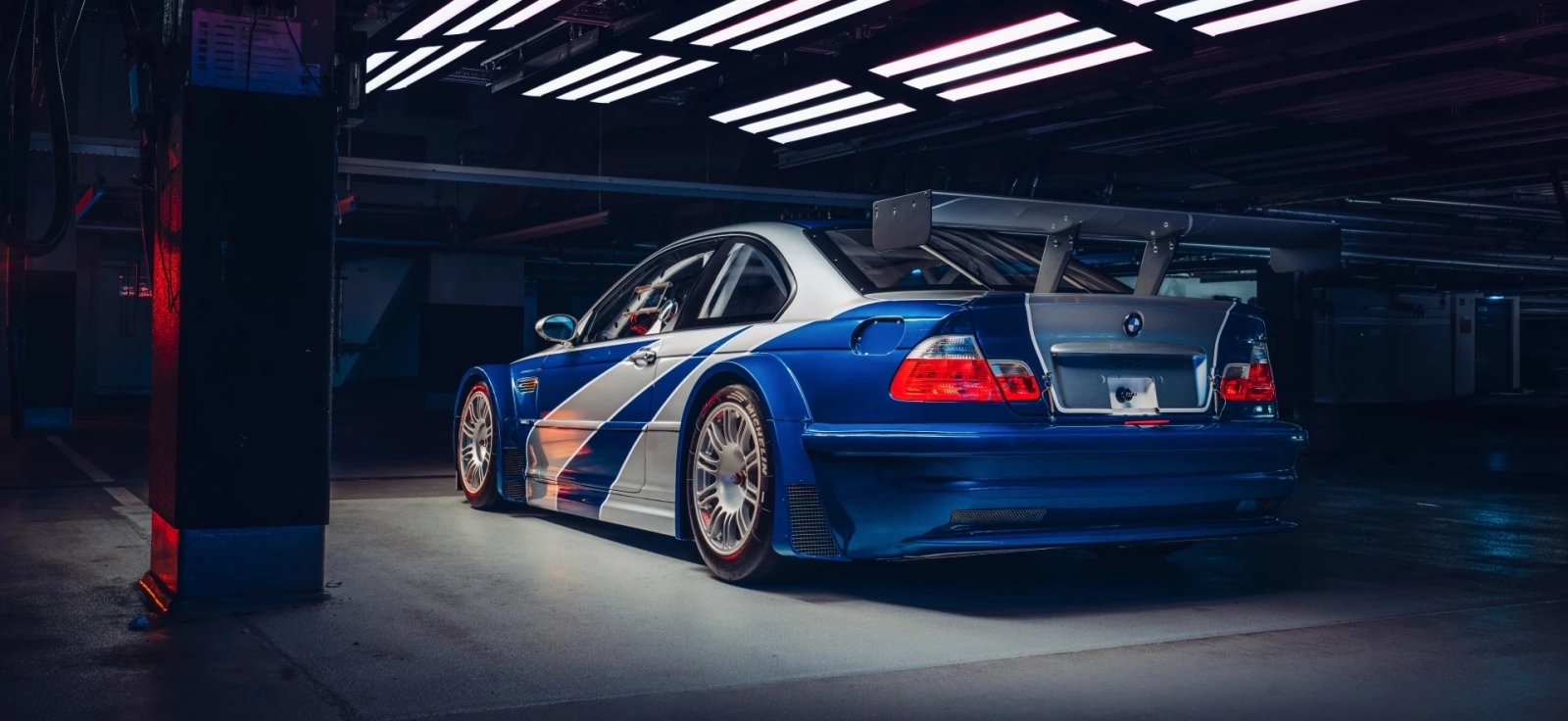 Bočni profil BMW M3 GTR sa prepoznatljivim plavim prugama i širokim lukovima točkova u tamnom garažnom okruženju