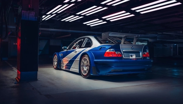 Bočni profil BMW M3 GTR sa prepoznatljivim plavim prugama i širokim lukovima točkova u tamnom garažnom okruženju