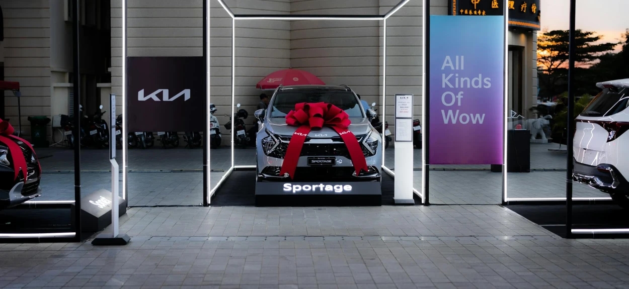 Automobil marke Kia parkiran u salonu, sa crvenom mašnom preko haube spreman za prodaju.