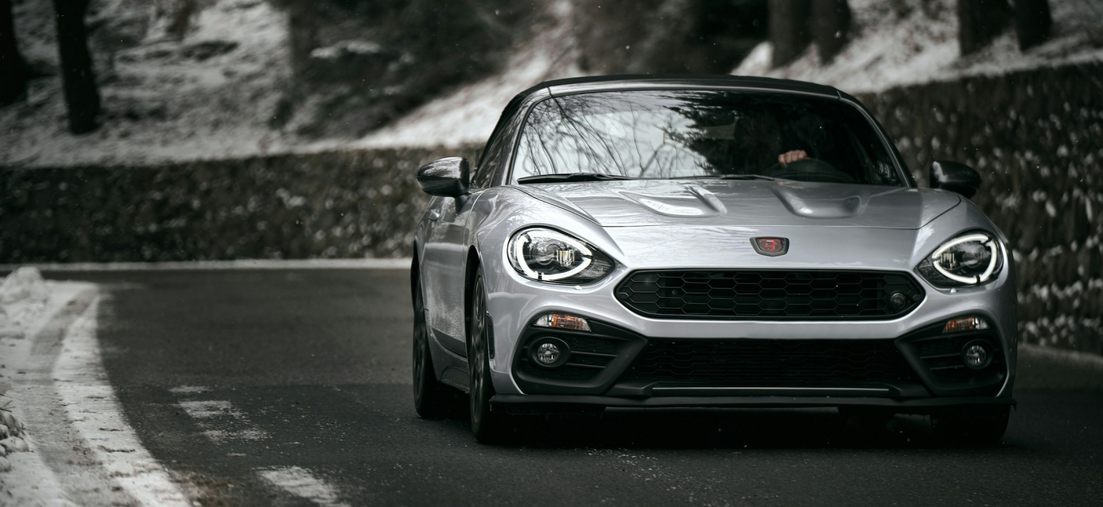 Prednji deo sivog sportskog automobila Abarth 124 Spider u vožnji na asfaltnom putu kroz šumu