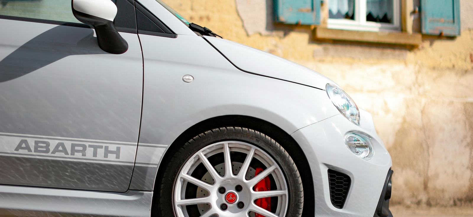 Bočni profil sivog Fiat Abarth sportskog automobila sa prepoznatljivom bočnom trakom i belim felnama parkiranog ispred zgrade