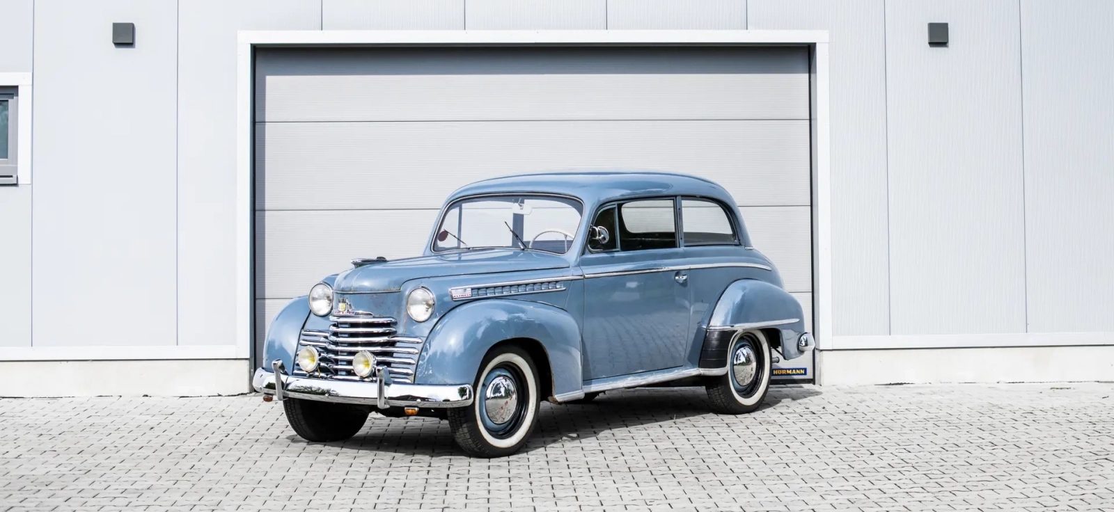 Klasican plavi automobil Opel Olympia sa samonosecom celicnom karoserijom parkiran ispred moderne bele garaze