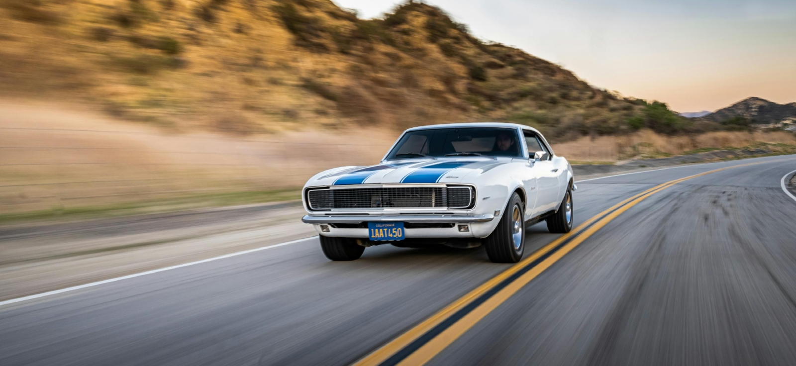 Beli Chevrolet Camaro prve generacije sa plavim trkačkim trakama u brzoj vožnji na otvorenom putu.