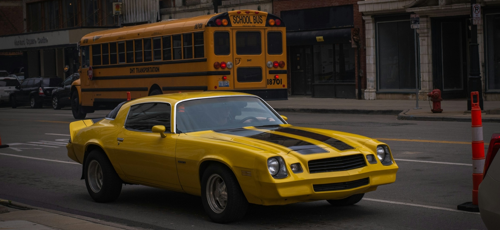 Klasični žuti Chevrolet Camaro druge generacije parkiran na gradskoj ulici ispred školskog autobusa.