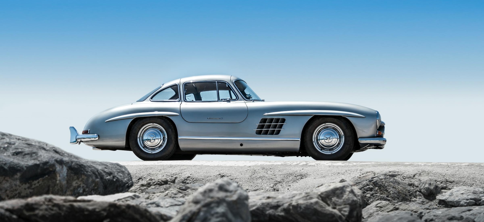 Profil srebrnog kupea Mercedes Benz 300SL na stenovitoj podlozi pod plavim nebom