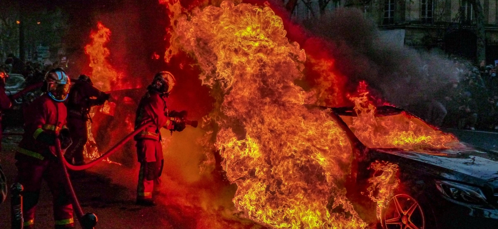 Auto koji gori i vatrogasni tim koji gasi požar.