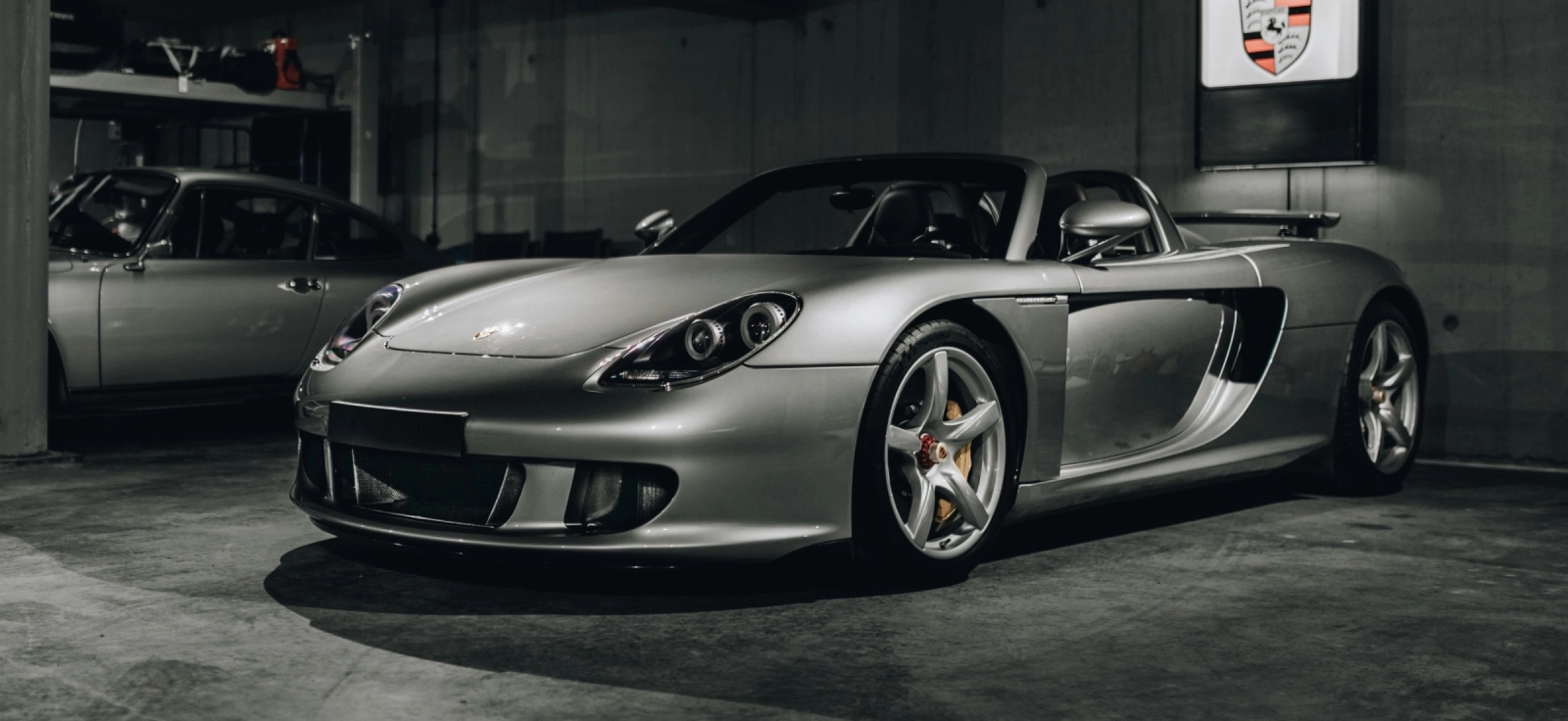 Porsche Carrera GT parkiran u garaži sa fokusom na prednji deo i prepoznatljiva prednja svetla ispod Porsche amblema na zidu