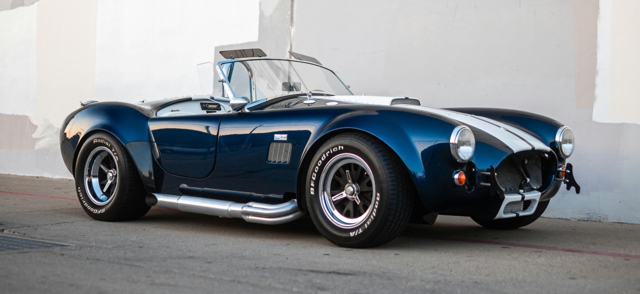 Legendarna plava Shelby Cobra 427 sa belim trakama parkirana pored zida, simbol američkog inata u auto-industriji