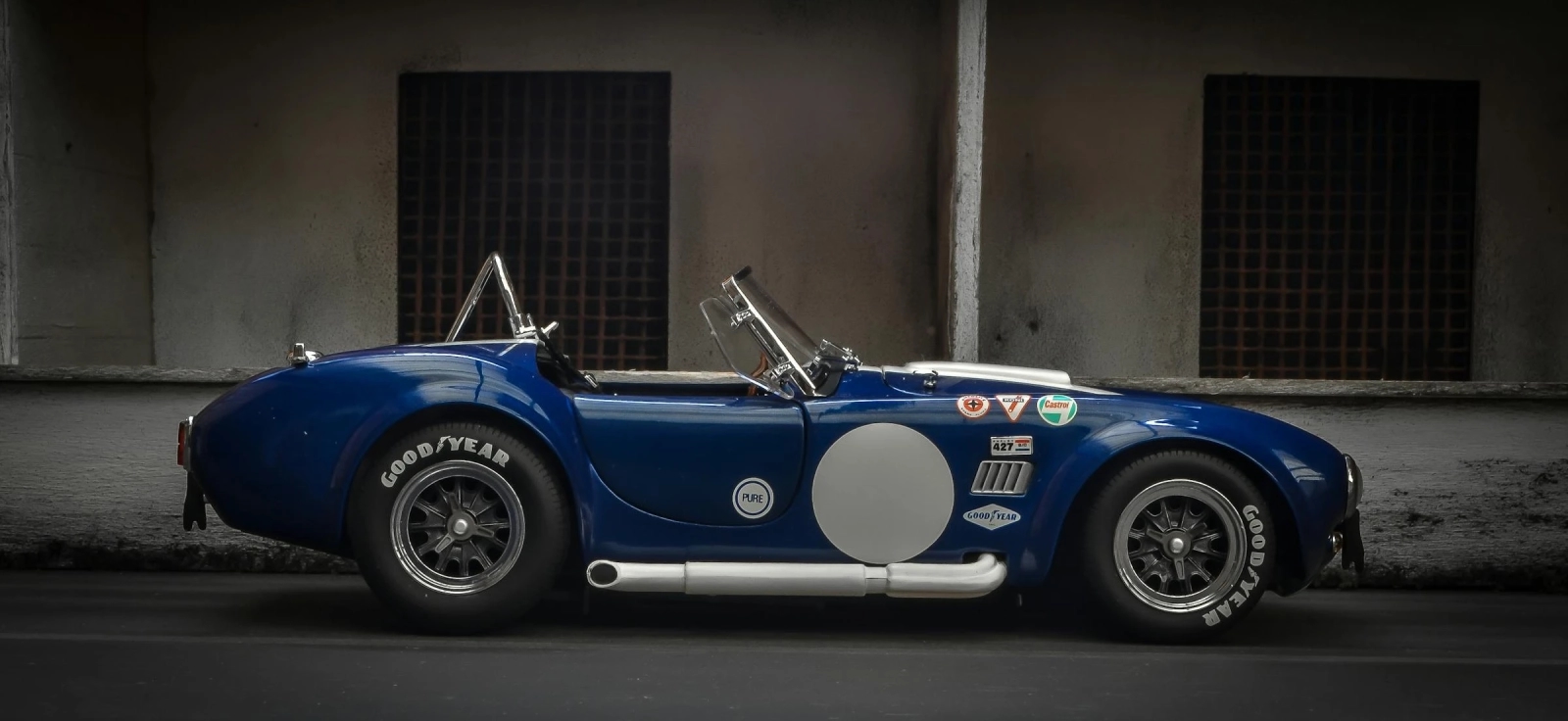 Bočna silueta plave Shelby Cobra 427 parkirane ispred industrijskog zida, klasični američki roadster sa britanskom šasijom