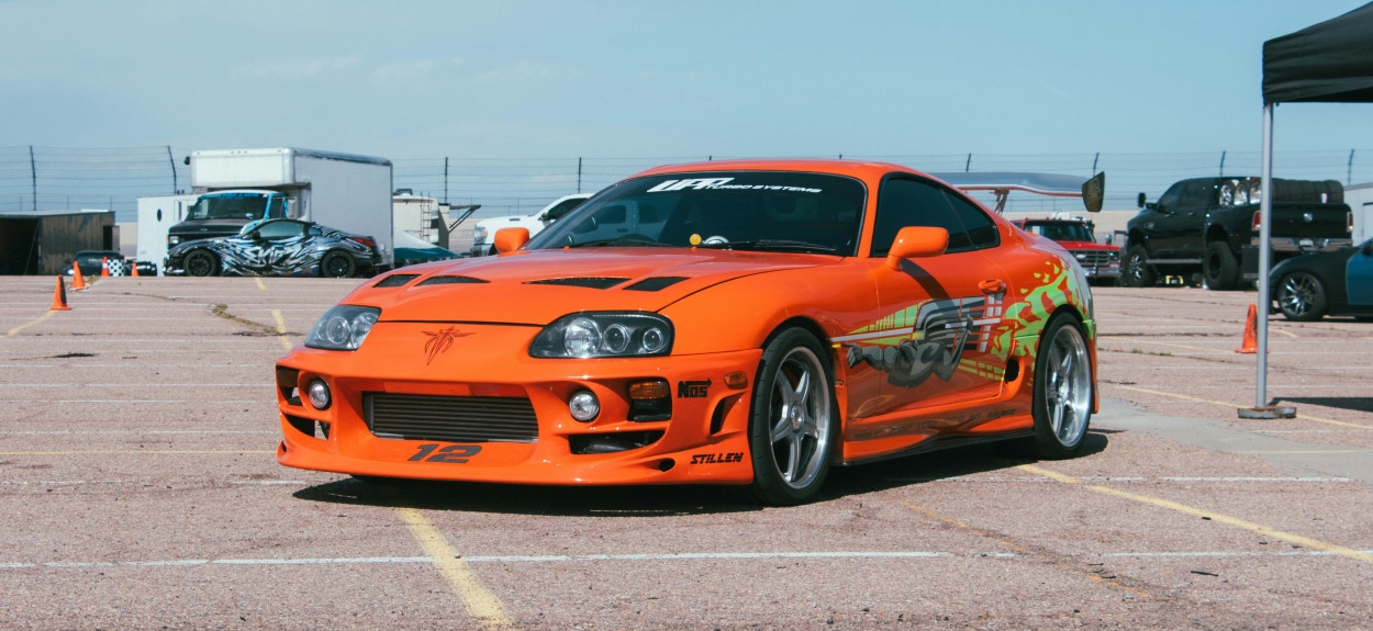 Narandžasta Toyota Supra MK4 sa grafikama inspirisana filmskim serijalom Fast and Furious (Paklene ulice), parkirana na skupu automobila