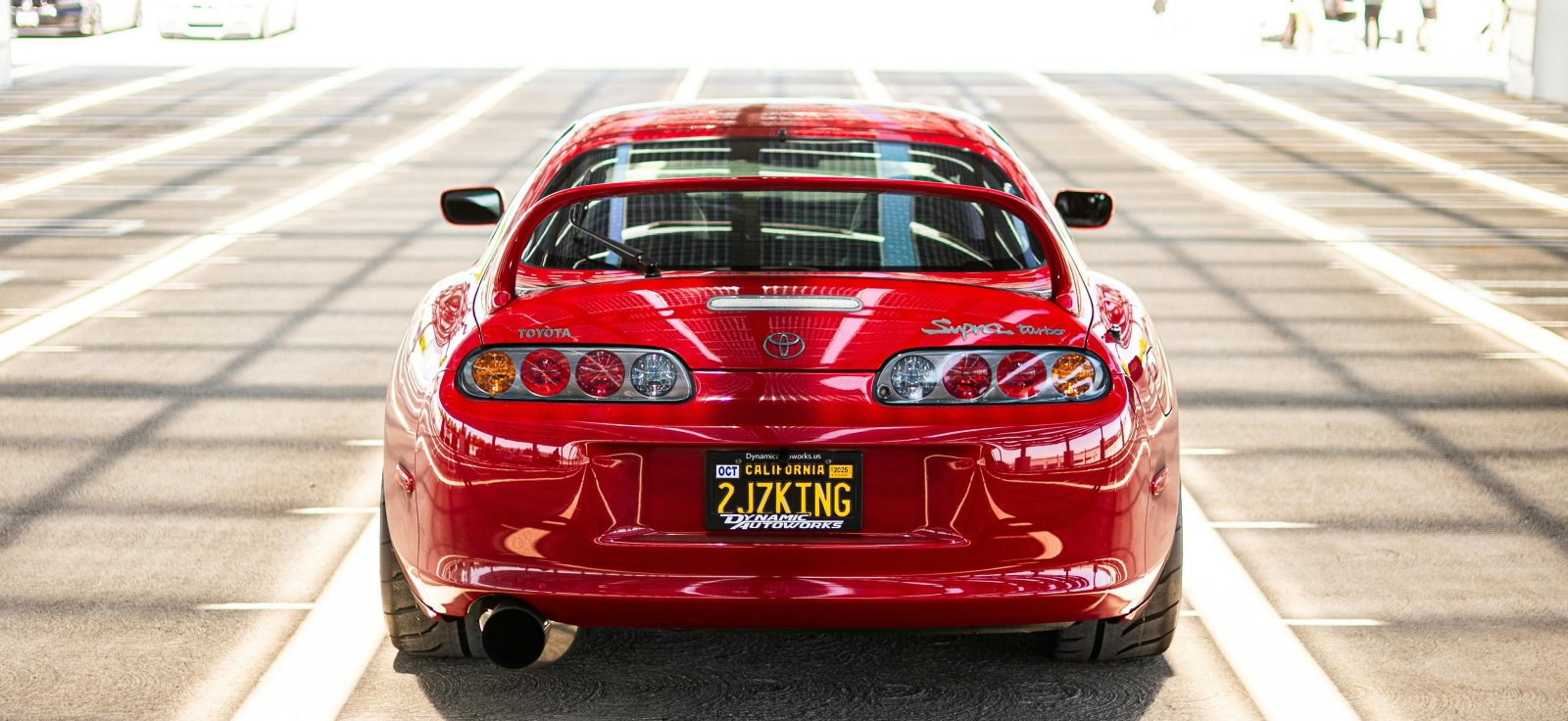 Pogled otpozadi na crvenu Toyota Supra MK4 sa velikim zadnjim spojlerom, parkiranu na krovu parkinga