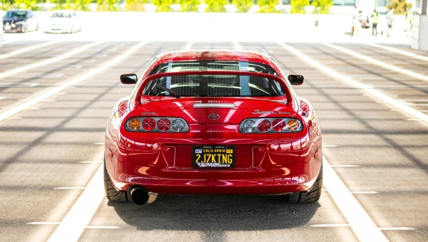 Pogled otpozadi na crvenu Toyota Supra MK4 sa velikim zadnjim spojlerom, parkiranu na krovu parkinga