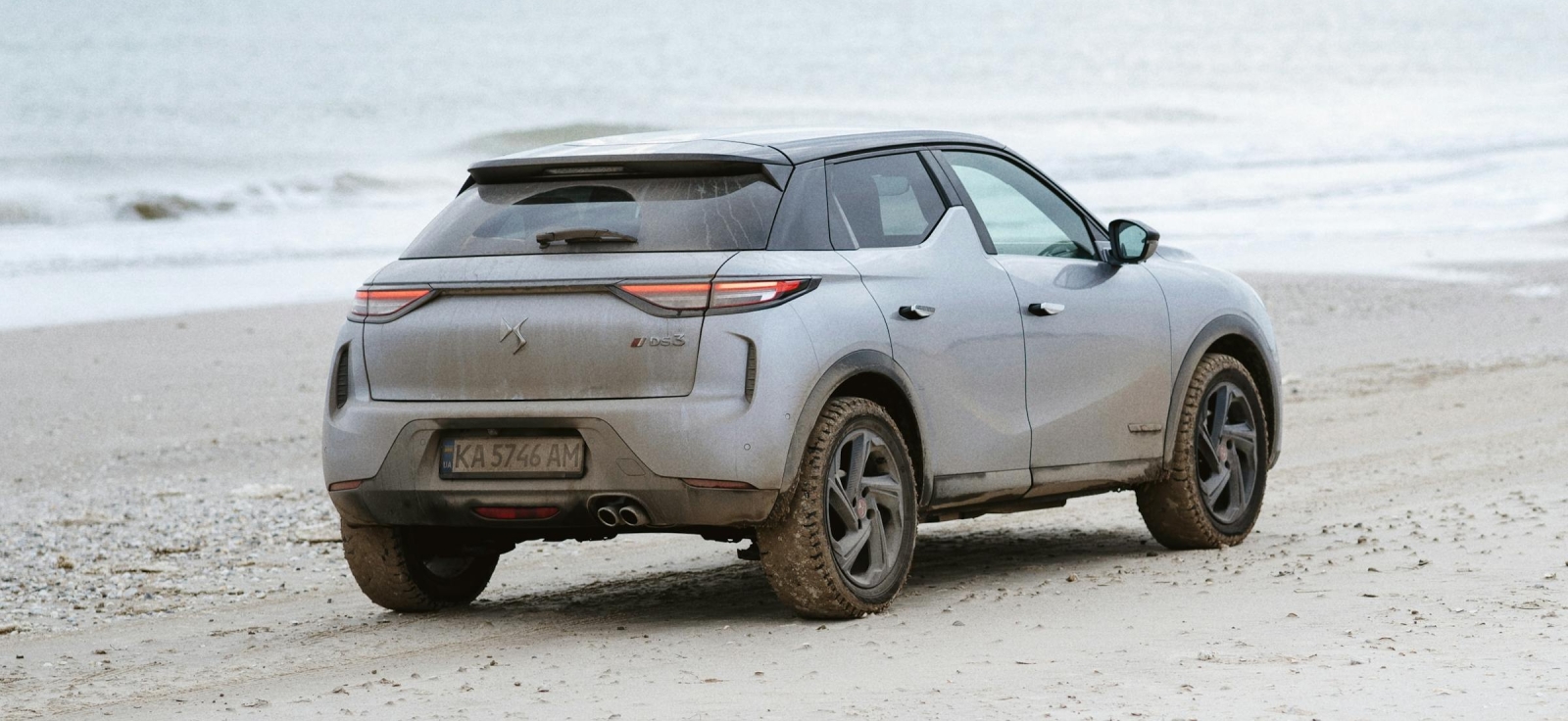 Sivi SUV automobil parkiran na peščanoj plaži pored mora pod oblačnim nebom